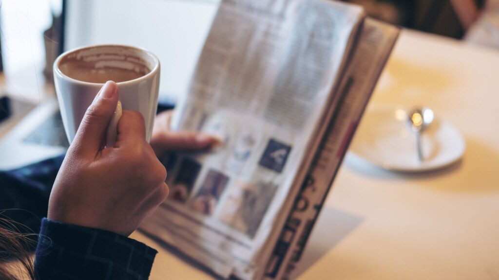 Person holding a white mug of coffee while reading a newspaper at a cafe table.