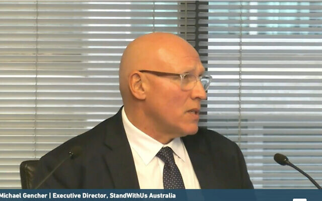 Profile view of a bald man in a dark suit and glasses speaking into a microphone during a meeting; a caption at the bottom lists a name and 'Executive Director, StandWithUs Australia'.