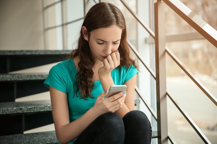 Upset teen girl sitting in a stairwell looking at her phone.
