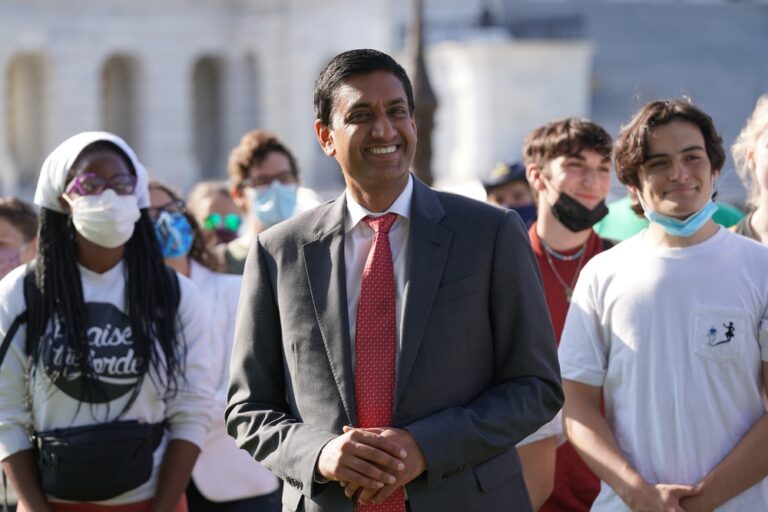 Smiling man in a gray suit and red tie stands at the center of a diverse crowd outdoors.