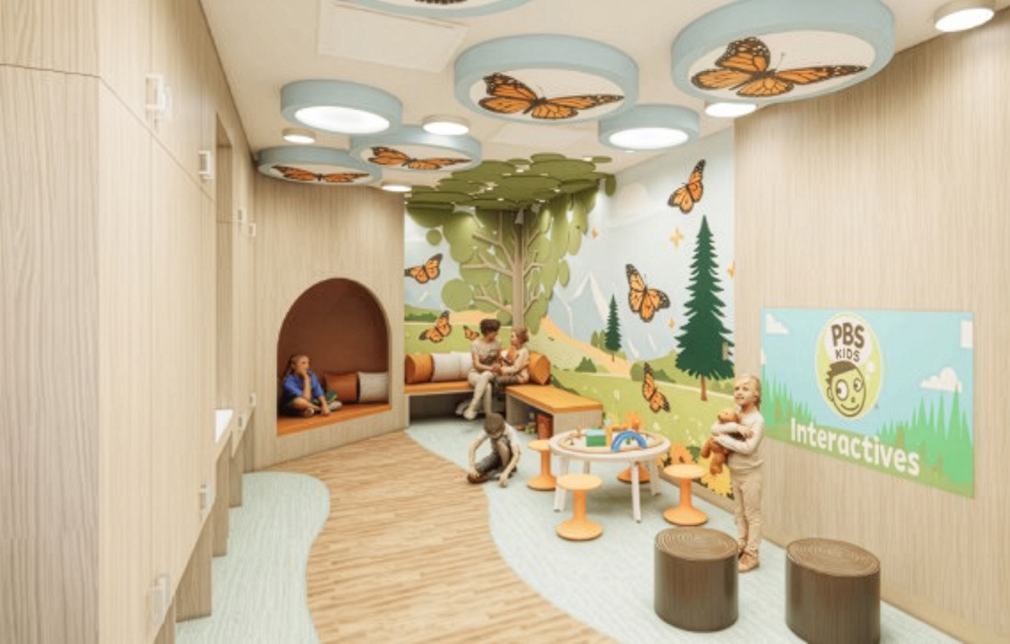Colorful children's playroom with a woodland mural, butterfly ceiling lights, and kid-sized seating for activities, labeled PBS Kids Interactives.