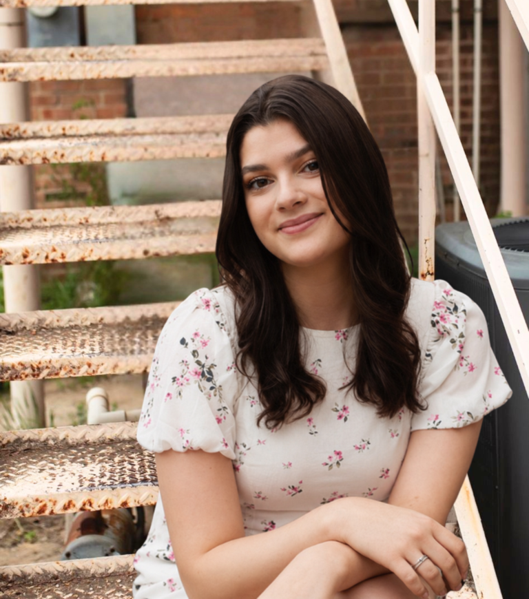 Young woman with long dark hair in a white floral dress sits on rusted outdoor metal stairs and smiles.