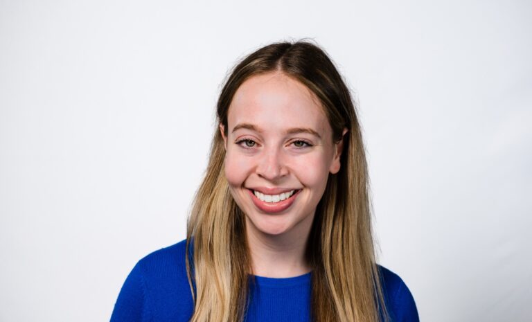 Smiling young woman with long light brown hair wearing a blue sweater, posed against a plain white background.