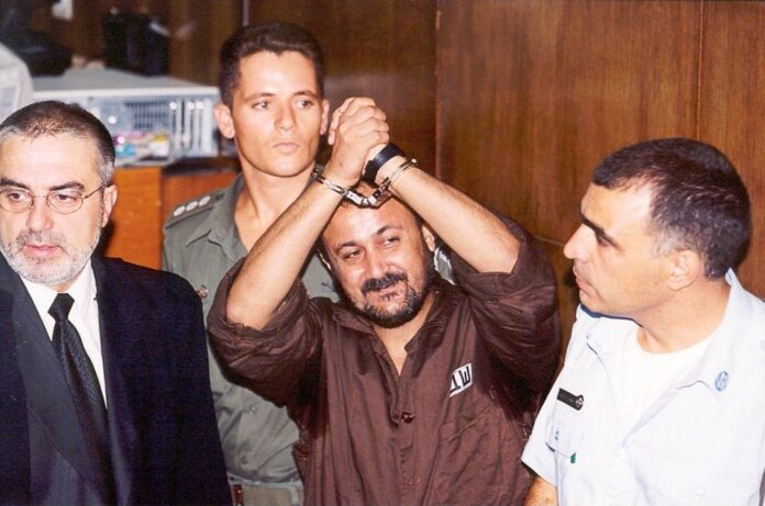 Man in a brown shirt with hands cuffed overhead, flanked by security personnel in a doorway or hallway setting