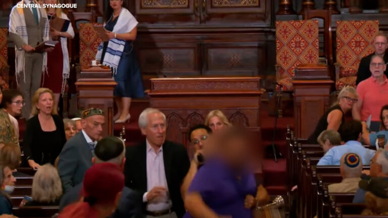 People mingle in a richly carved synagogue sanctuary, with ornate wooden pews and patterned chairs visible in the background.