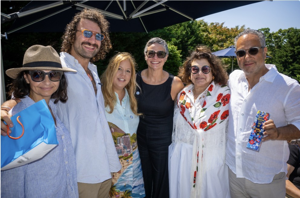 Six adults posing together outdoors at a sunny event, smiling for the camera under umbrellas.