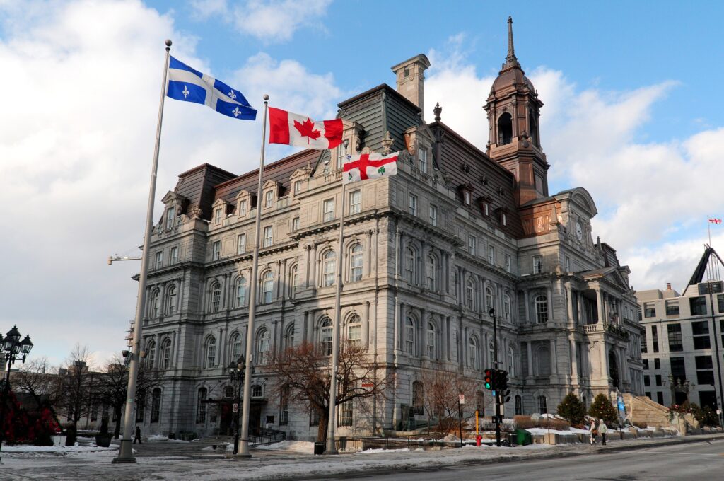 Historic stone city hall-style building with a tall tower and three flagpoles flying the Quebec, Canadian, and England flags against a blue sky.