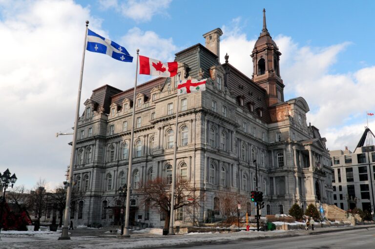 Historic stone city hall-style building with a tall tower and three flagpoles flying the Quebec, Canadian, and England flags against a blue sky.