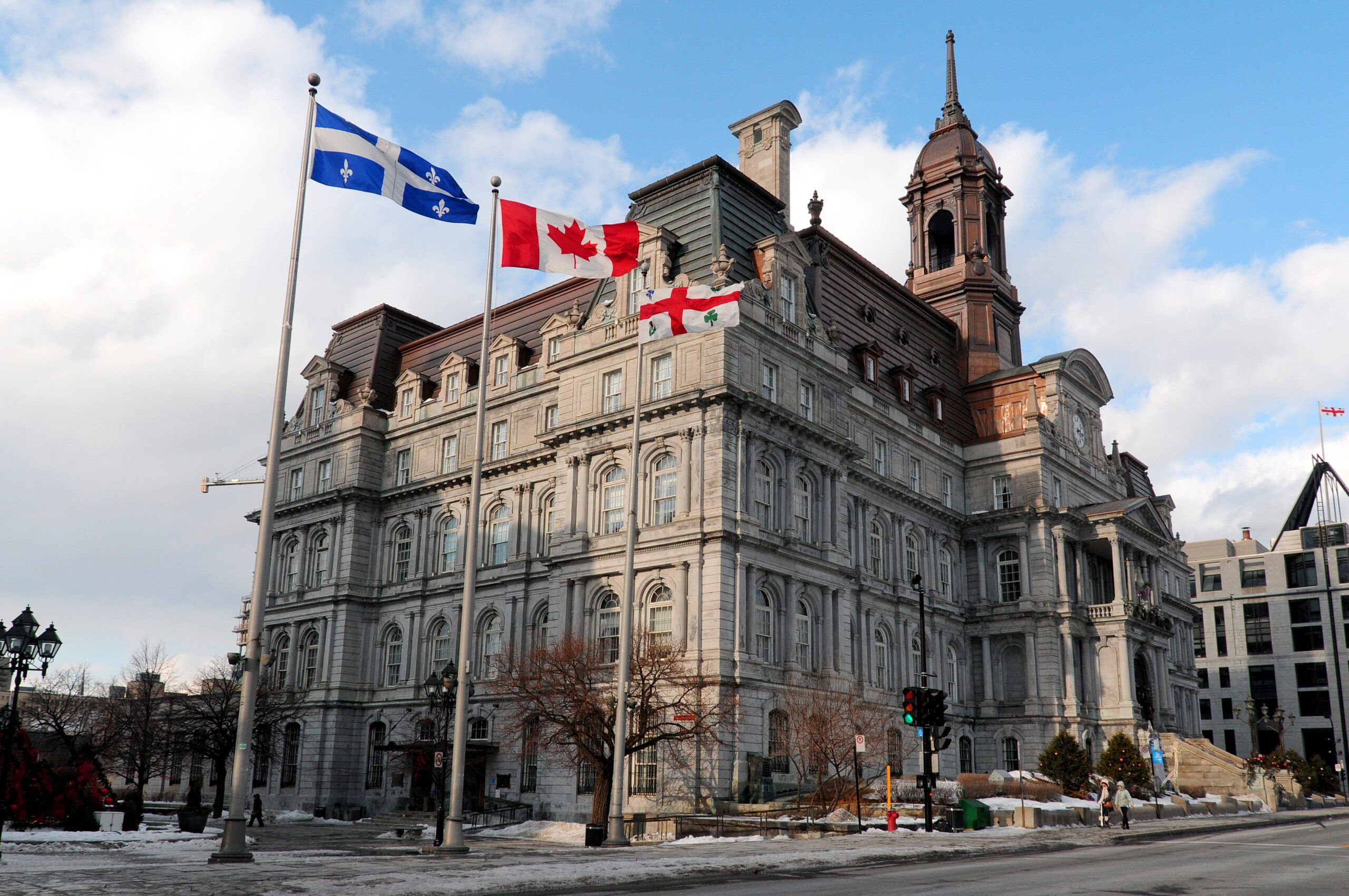 Historic stone city hall-style building with a tall tower and three flagpoles flying the Quebec, Canadian, and England flags against a blue sky.