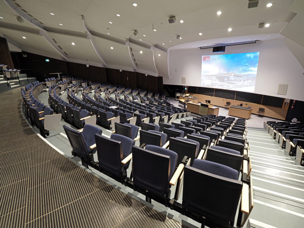 Lecture hall with curved rows of blue seats facing a large projection screen on the stage.