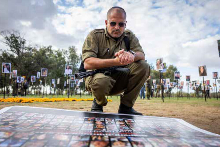 A uniformed man crouches over a large display of photos laid out on a table outdoors, with portrait boards on posts in the background at a memorial event.
