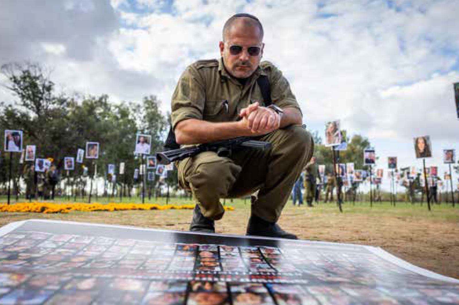 A uniformed man crouches over a large display of photos laid out on a table outdoors, with portrait boards on posts in the background at a memorial event.