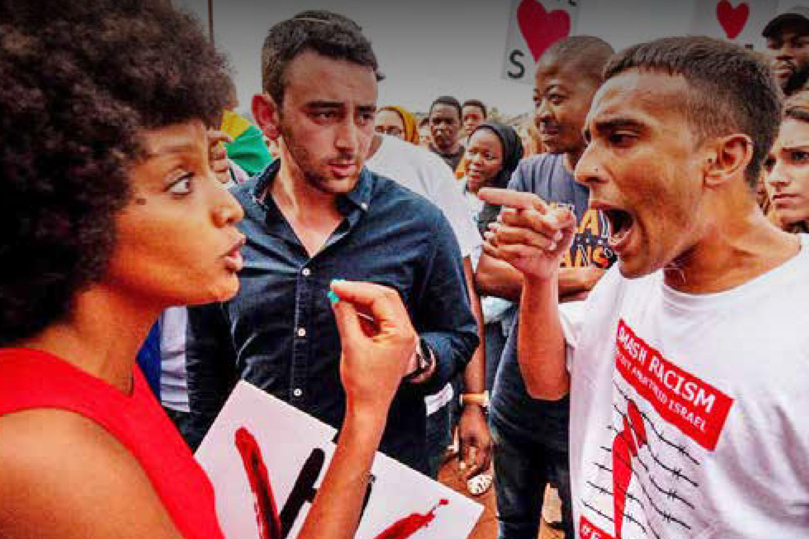 Diverse crowd at a protest; a woman in red speaks to a man while others hold signs about racism in the background.