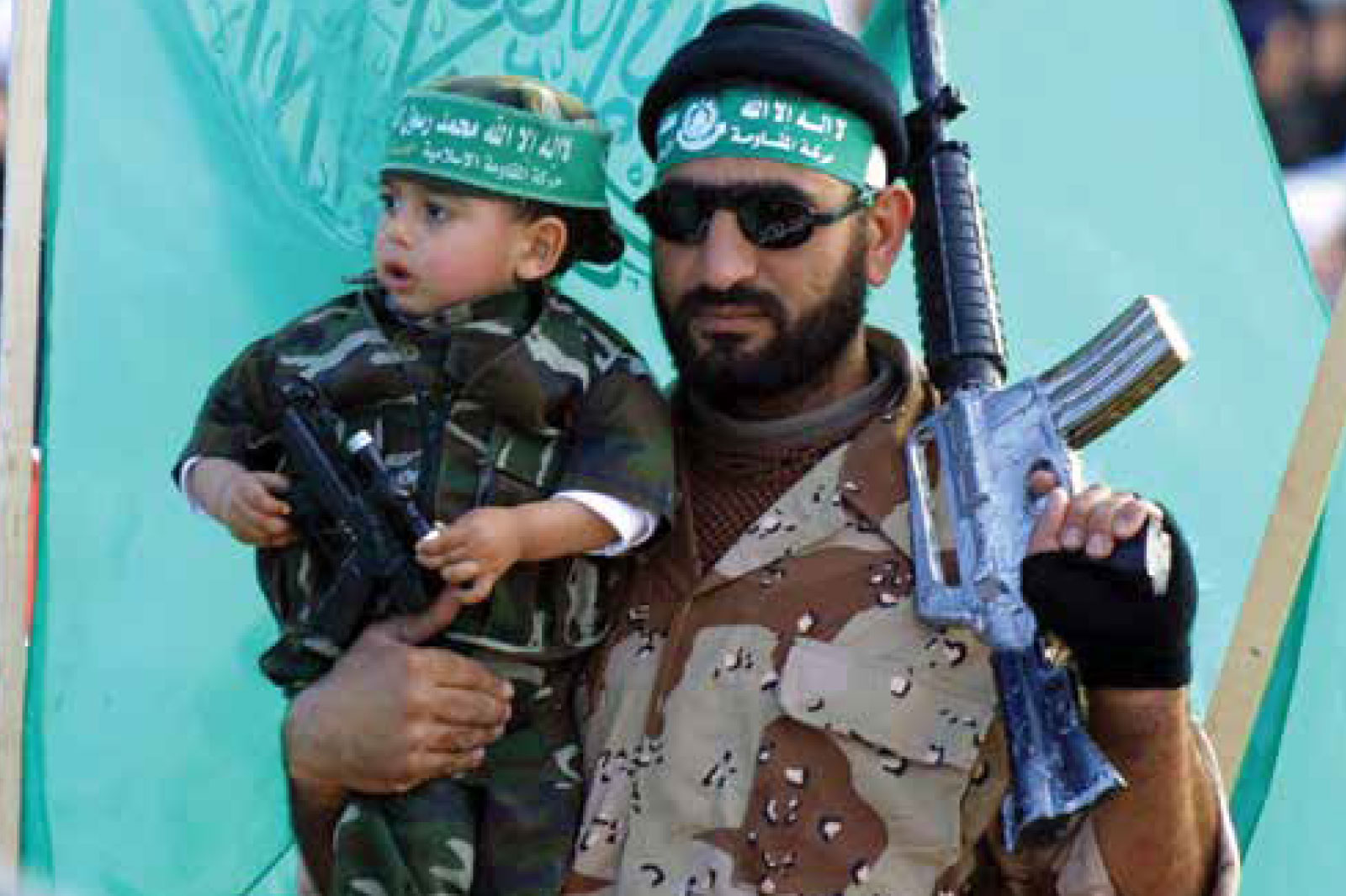 Bearded man in camo with a rifle and sunglasses, holding a young child wearing a matching green headband.